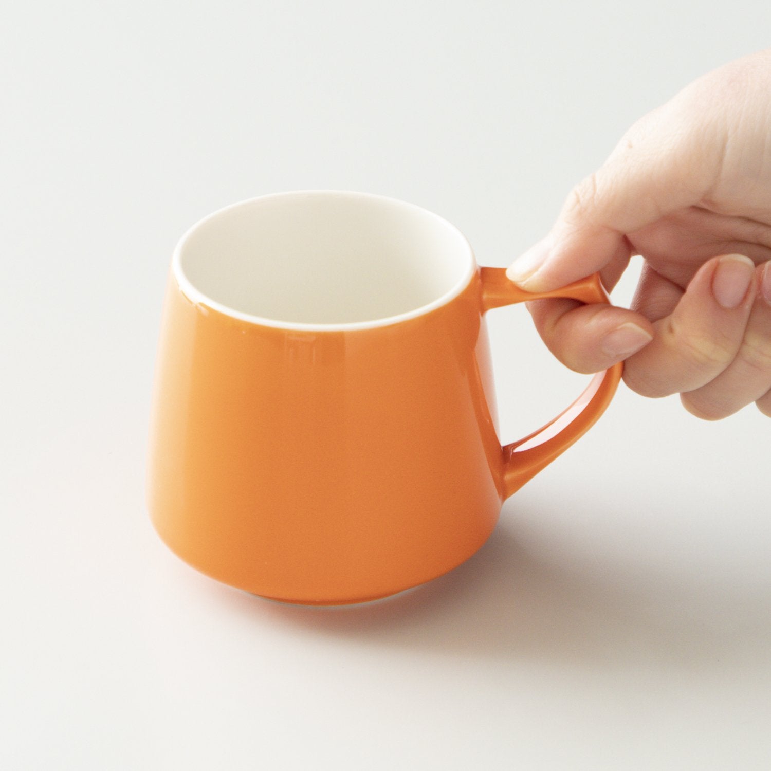 Orange mug with a hand holding its handle on a light gray background #orange